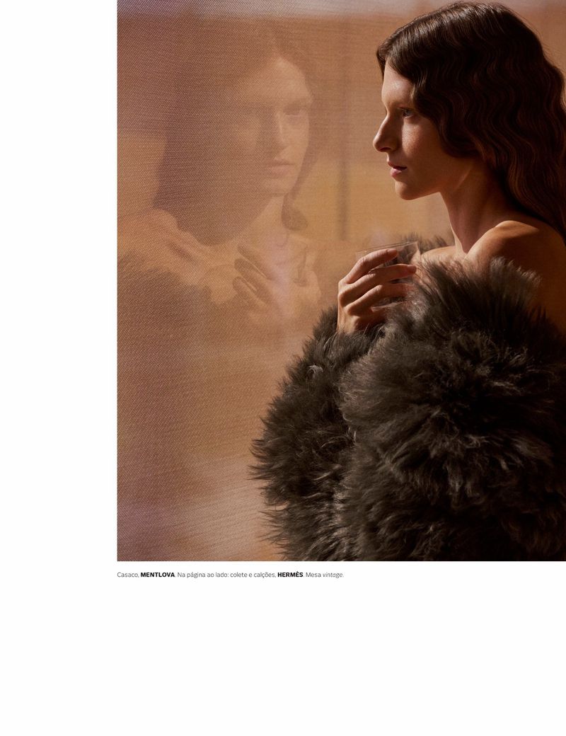 Fashion shot of a woman in profile wearing a fur coat with her reflection visible behind her in muted, diffused lighting.