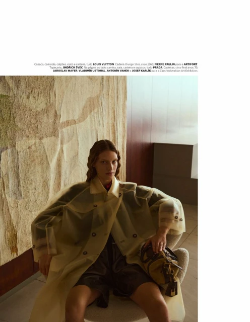 Fashion shot of a woman in a translucent trench coat, sitting on a chair, holding a duffel bag, with a patterned wall in the background.