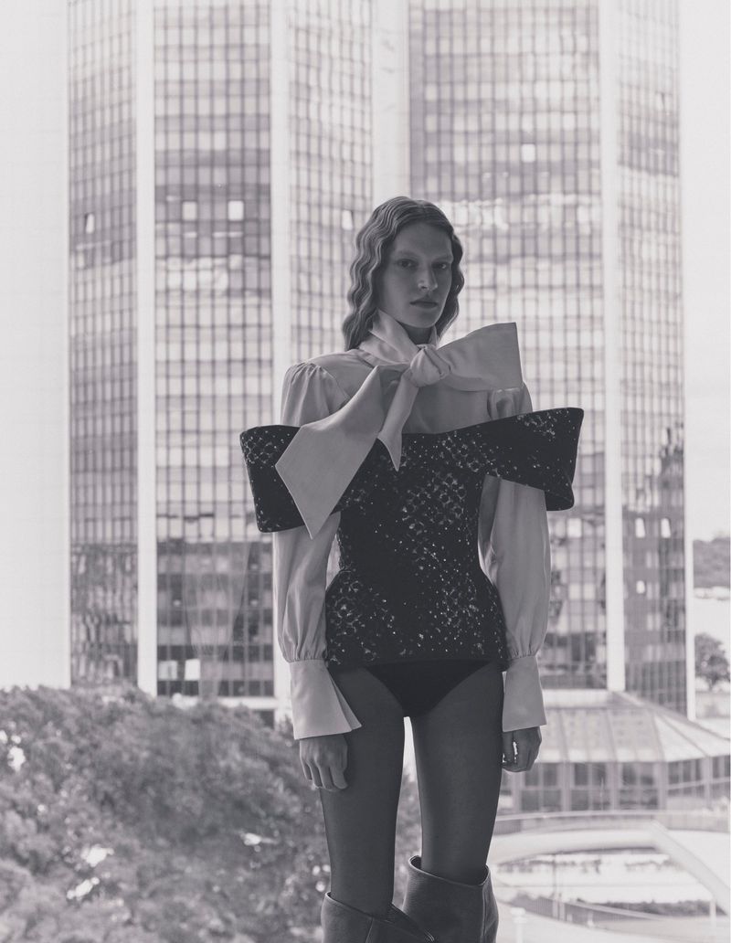 A woman stands in front of a window, wearing a structured top with a large bow, with a high-rise building visible in the background.