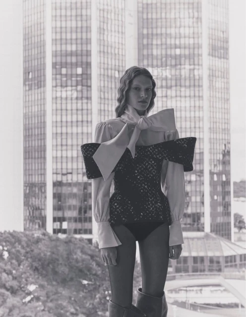 A woman stands in front of a window, wearing a structured top with a large bow, with a high-rise building visible in the background.