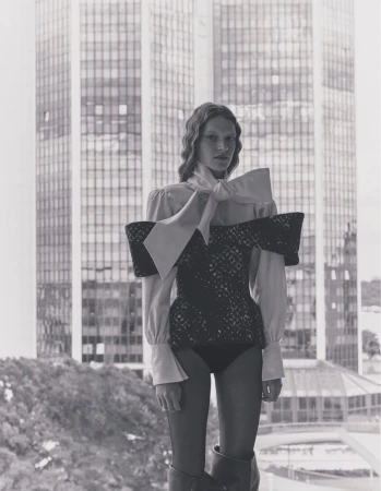 A woman stands in front of a window, wearing a structured top with a large bow, with a high-rise building visible in the background.