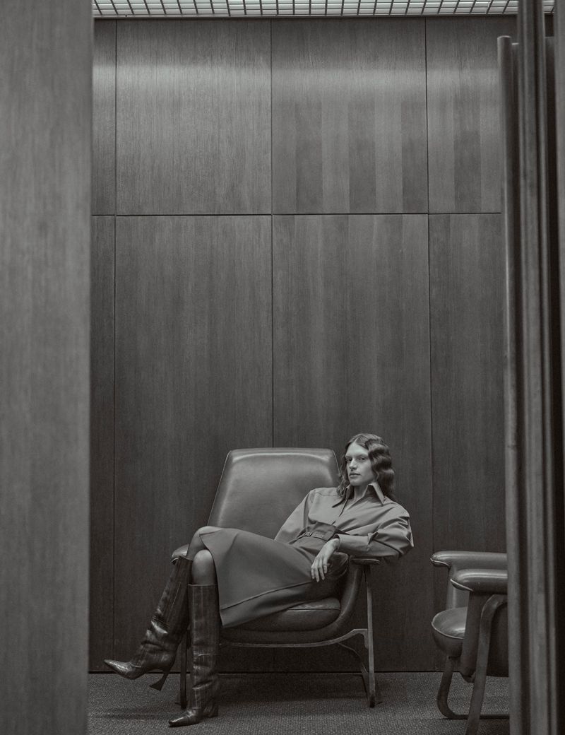 A woman sits in a leather chair in a monochromatic indoor space, wearing a long-sleeved dress and knee-high boots, showcasing fashion.