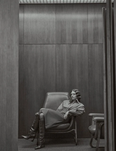 A woman sits in a leather chair in a monochromatic indoor space, wearing a long-sleeved dress and knee-high boots, showcasing fashion.