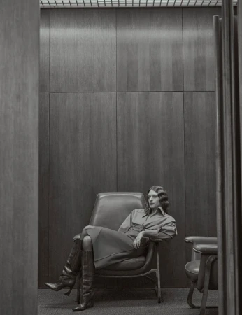 A woman sits in a leather chair in a monochromatic indoor space, wearing a long-sleeved dress and knee-high boots, showcasing fashion.
