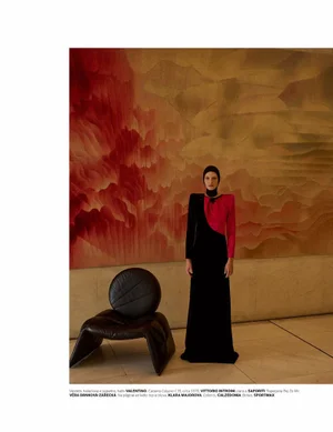 Model wearing a black and red dress with a headpiece standing next to a black leather chair in front of a textured wall