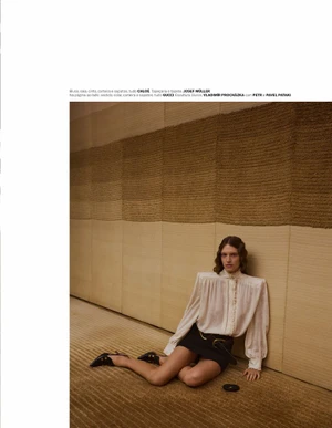 Model wearing a white blouse and black skirt seated on a textured carpet against a paneled wall for Vogue Portugal