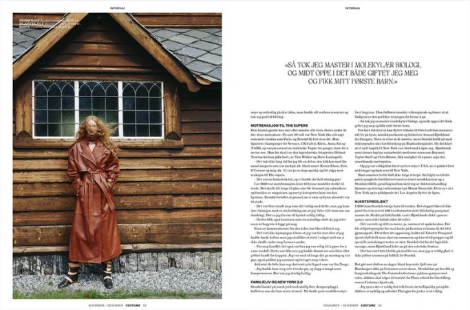 Editorial fashion shot of a redhead woman in a brown leather trench coat posing outdoors against a rustic wooden house with window.