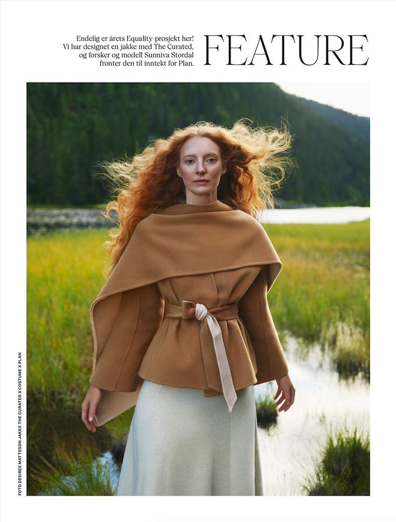 A woman with long, red hair wears a stylish belted tan coat in a grassy field with a lake and mountain backdrop, fashion editorial. 