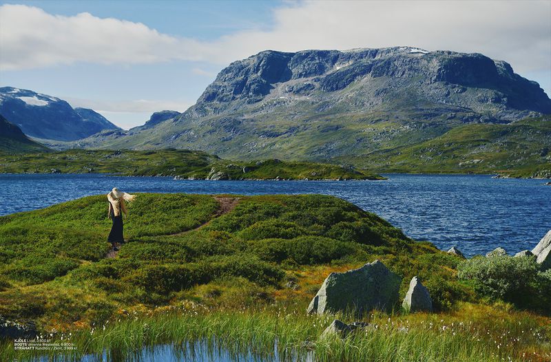 A woman in a black dress and straw hat walks on a grassy hill overlooking a scenic lake with large mountains in the background on a sunny day.