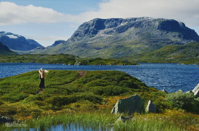 A woman in a black dress and straw hat walks on a grassy hill overlooking a scenic lake with large mountains in the background on a sunny day.