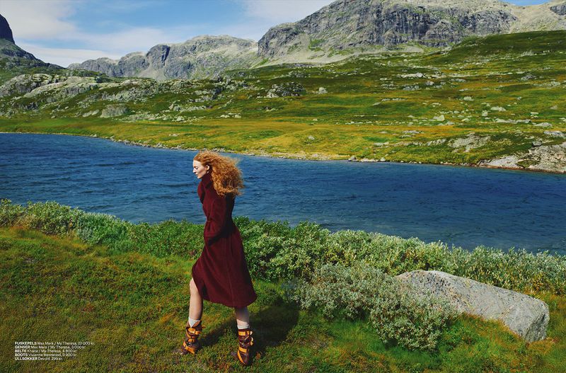 A red-haired woman in a maroon coat walks along a grassy field by a blue lake, with mountains and a clear sky in the background.