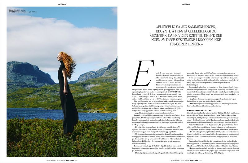 Woman in a long black dress stands in a grassy field against a backdrop of mountains and a blue lake on a partly cloudy day.