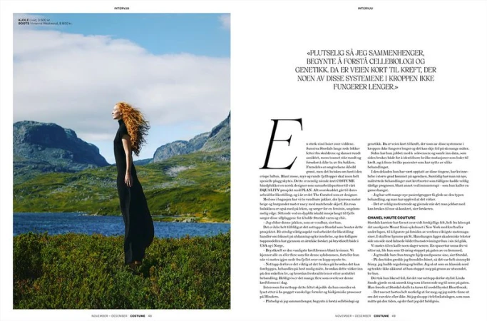 Woman in a long black dress stands in a grassy field against a backdrop of mountains and a blue lake on a partly cloudy day.