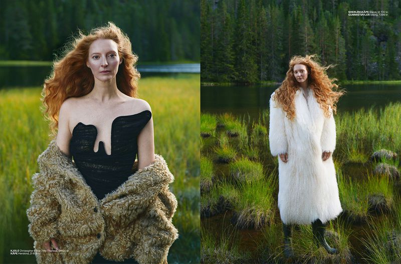 Fashion editorial shot of a redhead model in a black dress and fur coat standing outdoors in a nature setting by a lake.