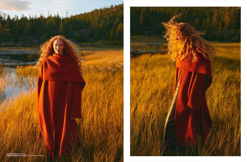 A long-haired woman in an orange poncho stands in a field of tall golden grass near a lake, with a forest visible in the background.