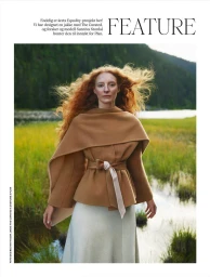 Red-haired model wearing a camel wrap jacket and light skirt outdoors in a grassy landscape with a river and hills.