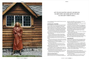 Editorial double-page spread featuring a model in a long brown leather coat standing beside a wooden building.
