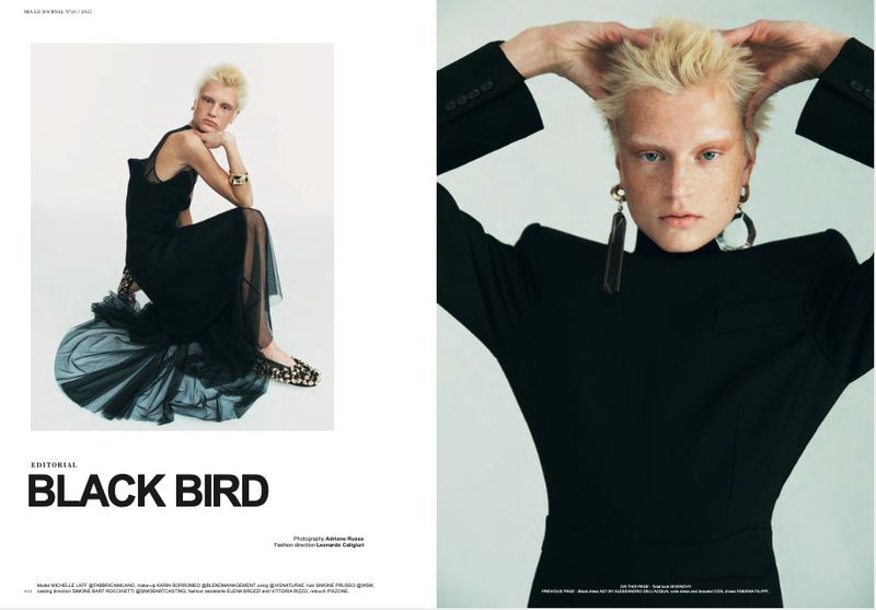 Editorial fashion shot: a blonde woman in a black dress poses indoors against a white backdrop, complemented by geometric jewelry and sleek footwear.