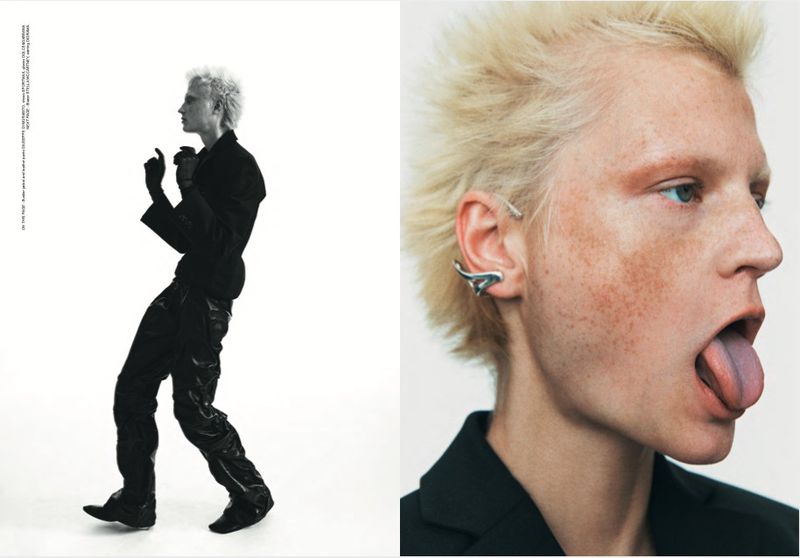 A fair-skinned person with blond hair wears a black jacket and strikes a pose against a white backdrop in a fashion editorial-style shot.