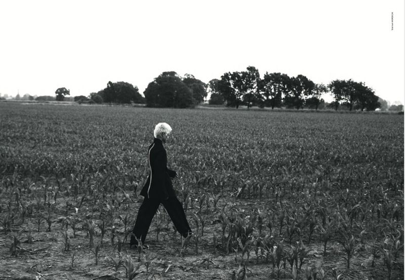 Monochrome portrait of an adult with short hair, walking through a field, wearing a dark suit with trees in the background.