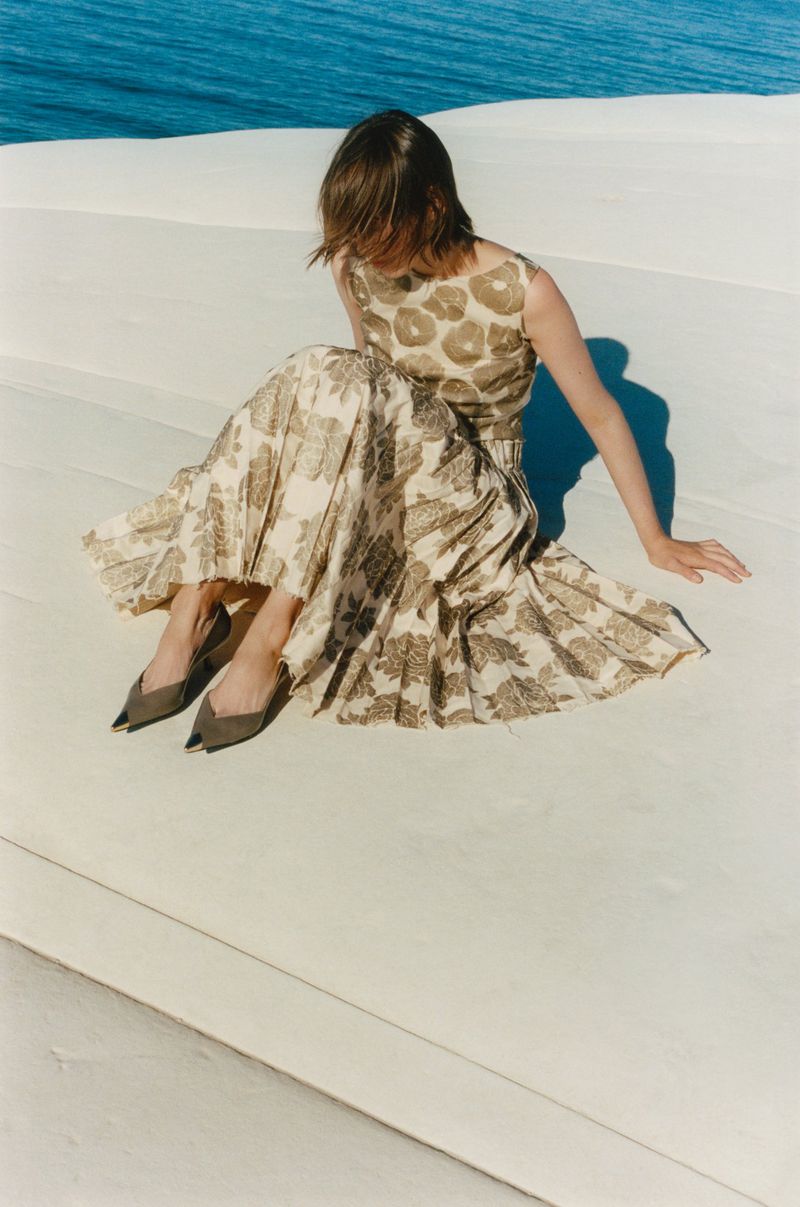 Model Thea Almqvist seated on a sloping white rock wearing a patterned beige sleeveless top, pleated skirt, and pointed heels by the sea.