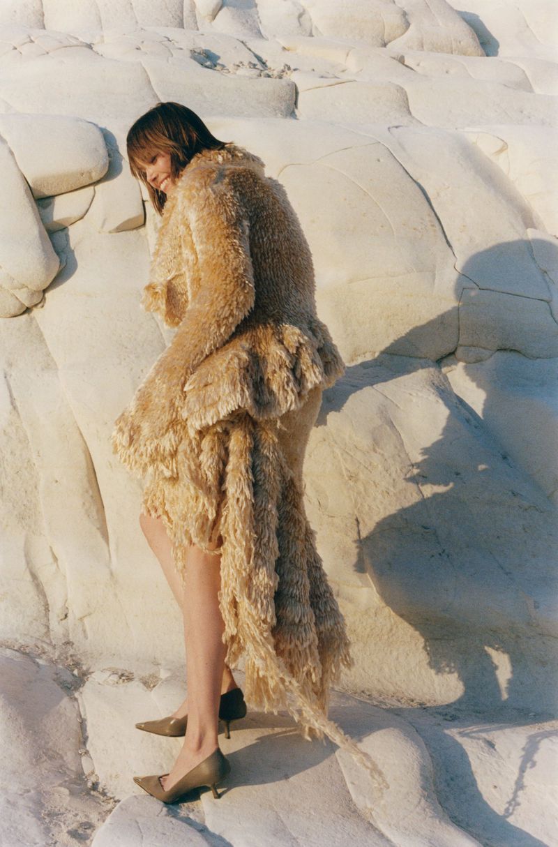 Model wearing a textured beige fringe coat and heels standing against white rocky cliffs