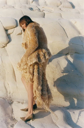 Model wearing a textured beige fringe coat and heels standing against white rocky cliffs