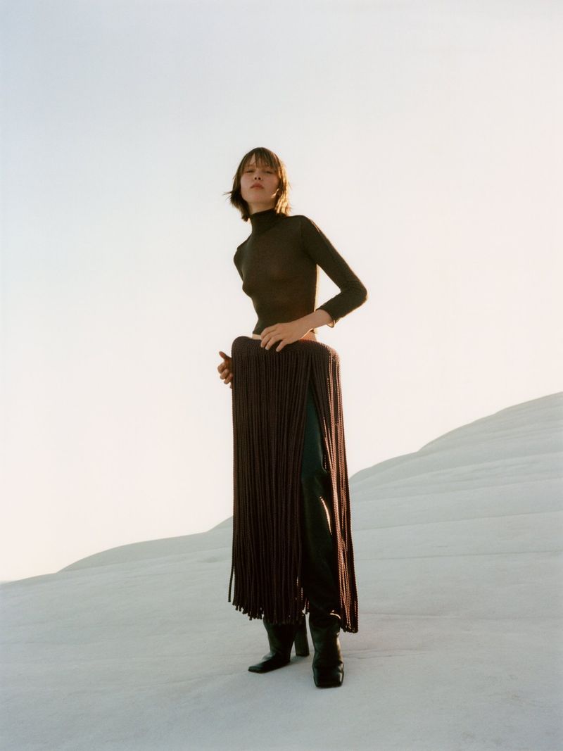 A woman stands in a minimalist landscape, wearing a brown turtleneck, black leather pants, boots, and a long, fringed brown cape.