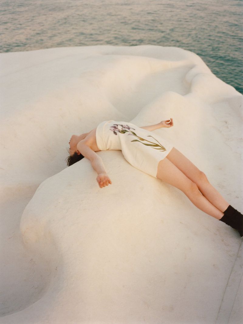 A woman in a white dress with a floral design is lying on white rock formation, with the blue sea visible in the background.