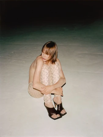 Model Thea Almqvist seated on a pale surface wearing patterned tights and black mules in a project by Jeremy Everett