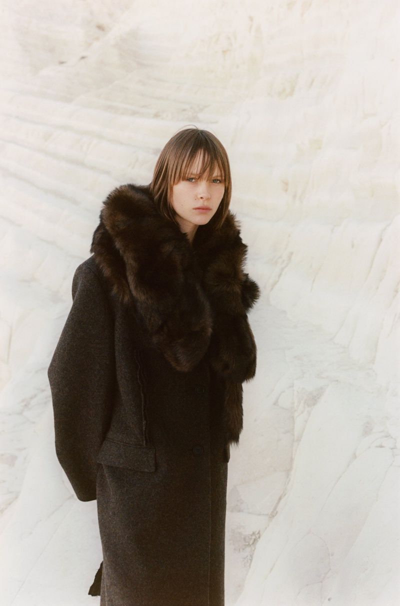 Woman in dark winter coat with fur collar poses against a white textured background, captured in a fashion-forward medium shot.