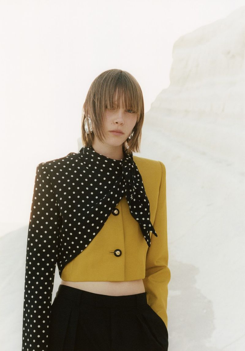A slender woman with blunt bangs poses in a color-blocked yellow and black polka dot blazer and black pants against a white background.