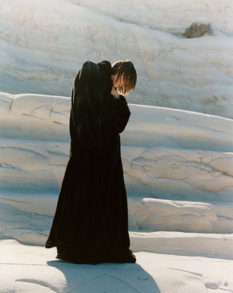 A person in a black robe stands on a white stone landscape with head bowed, bathed in sunlight, creating a dramatic yet serene scene.