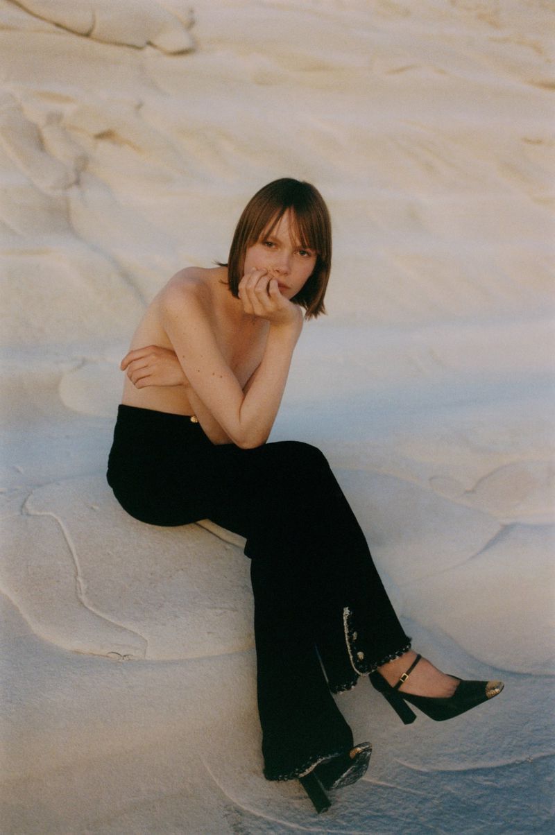 Brunette woman in black pants poses sitting on a white rock formation in nature, wearing heels and facing the camera.
