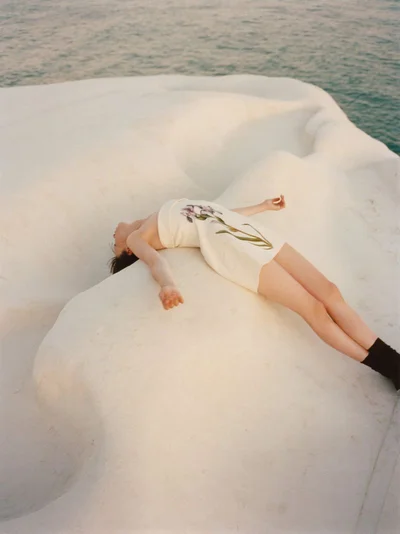 Model lying on white rock formation beside the sea wearing a white mini dress with floral print