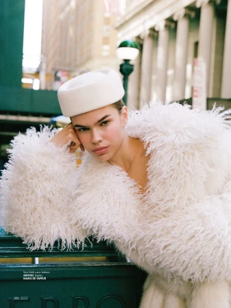 Woman in a white furry coat and hat leans against a green metal structure in an urban environment with tall buildings.