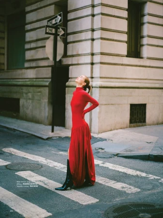 A young adult female model in a red dress and black boots poses on a crosswalk in an urban setting with a light brick building.