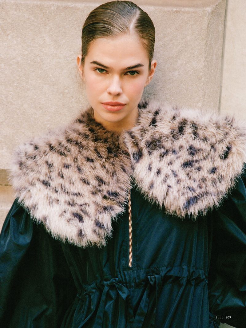 A portrait of a young adult woman wearing a dark blue jacket with a cheetah print collar against a neutral wall backdrop.