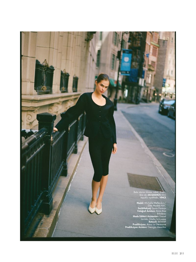 Fashion shot of a woman in a black pantsuit and cream heels walking on a city sidewalk, buildings in the background.
