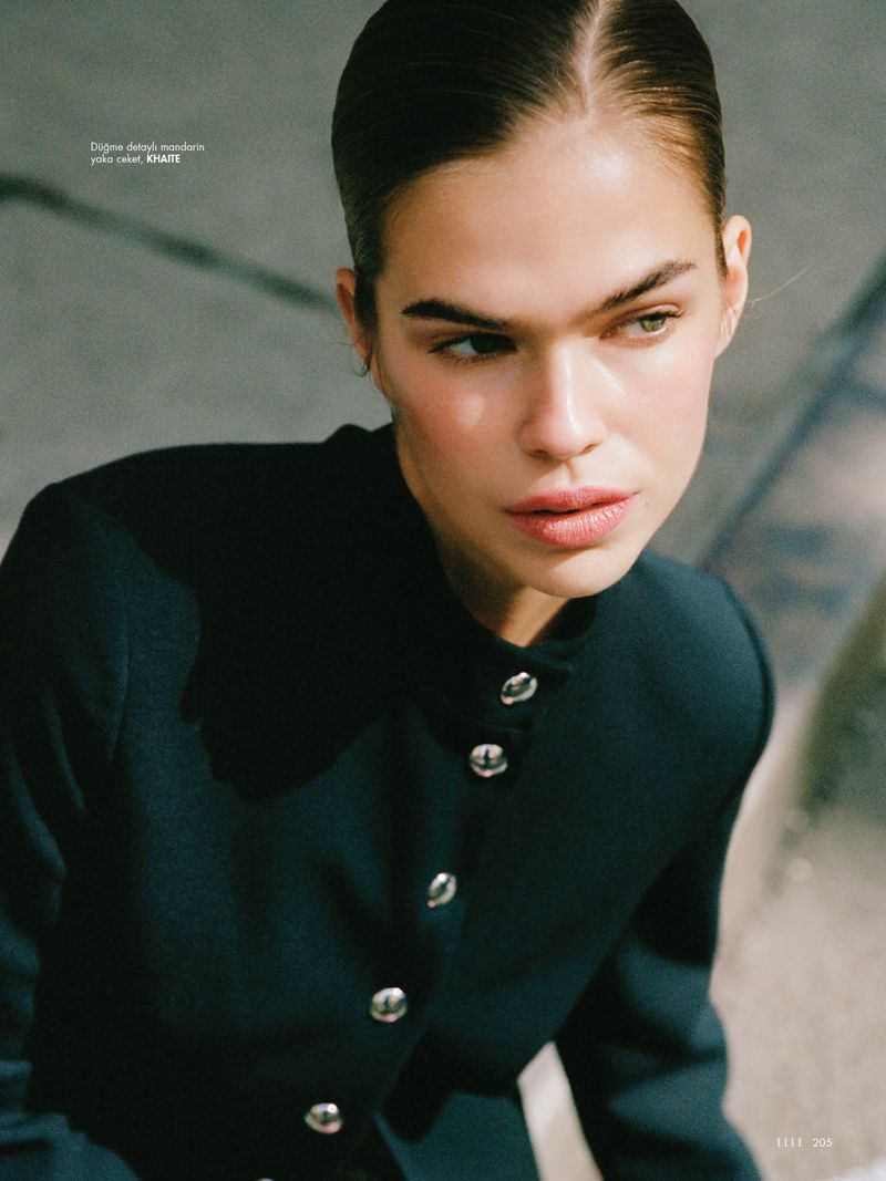 Close-up of a poised adult woman with slicked-back hair and dark coat, set against a muted background in a beauty and fashion editorial style.
