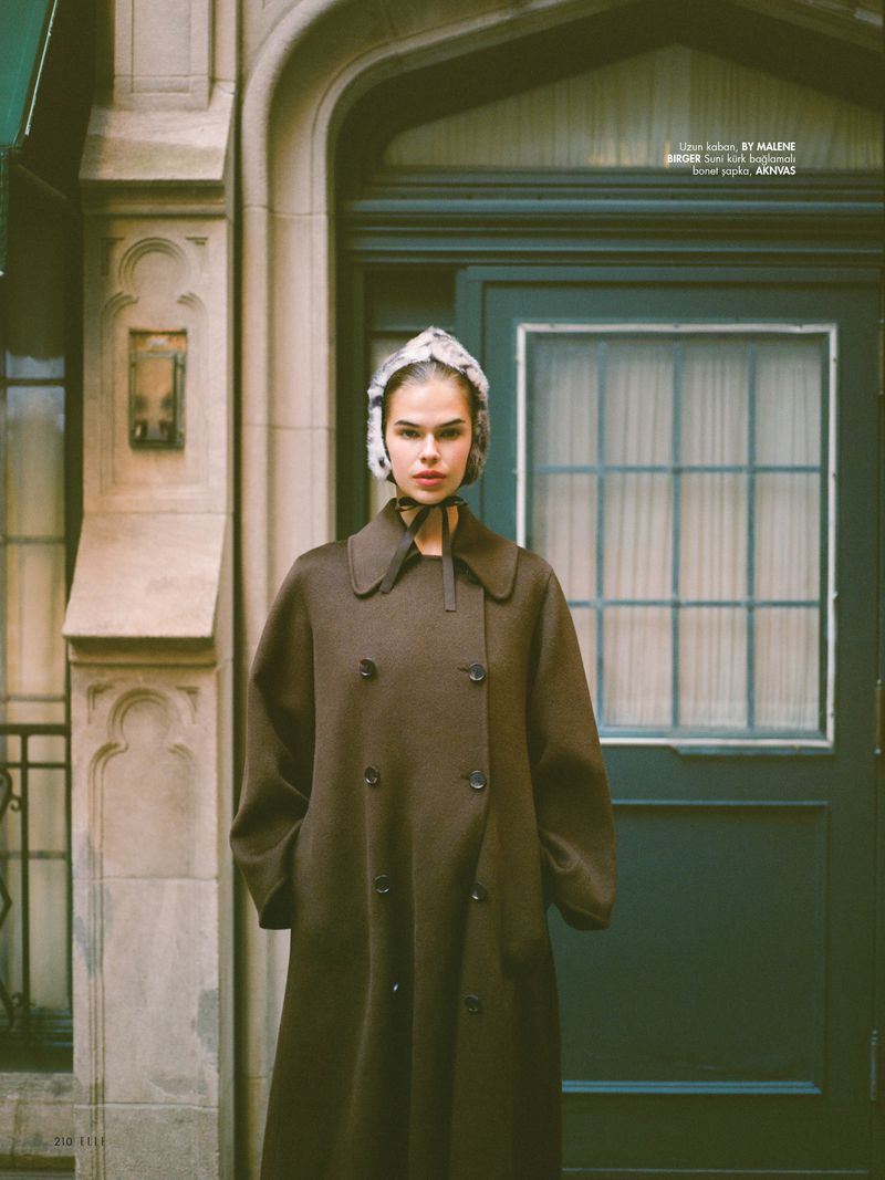 A woman wearing a brown coat and furry hat stands in front of a green door in an outdoor urban setting.