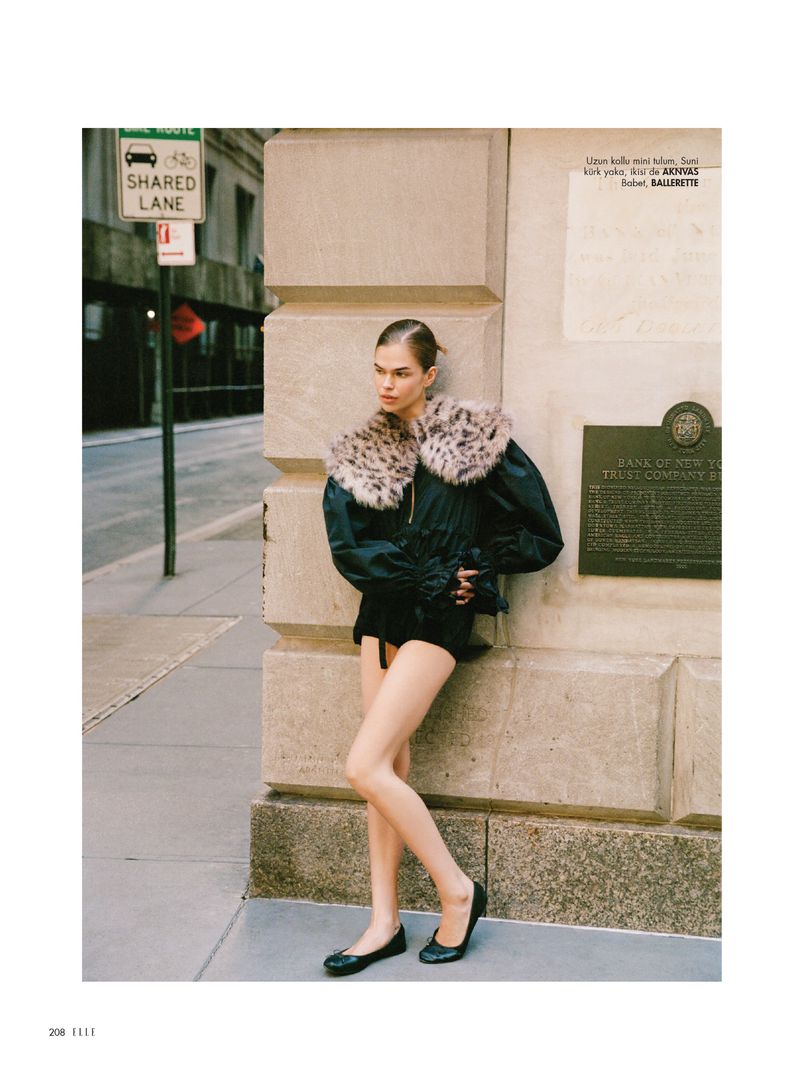 Fashion editorial shot in urban setting featuring a female model wearing a black outfit with a fur collar leaning against a building.