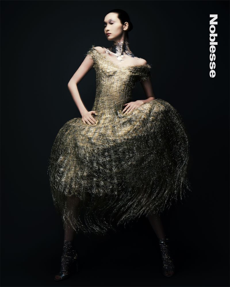 Elegant Asian woman in a gold wire gown and metallic heels poses against a dark backdrop, creating a striking fashion editorial image.