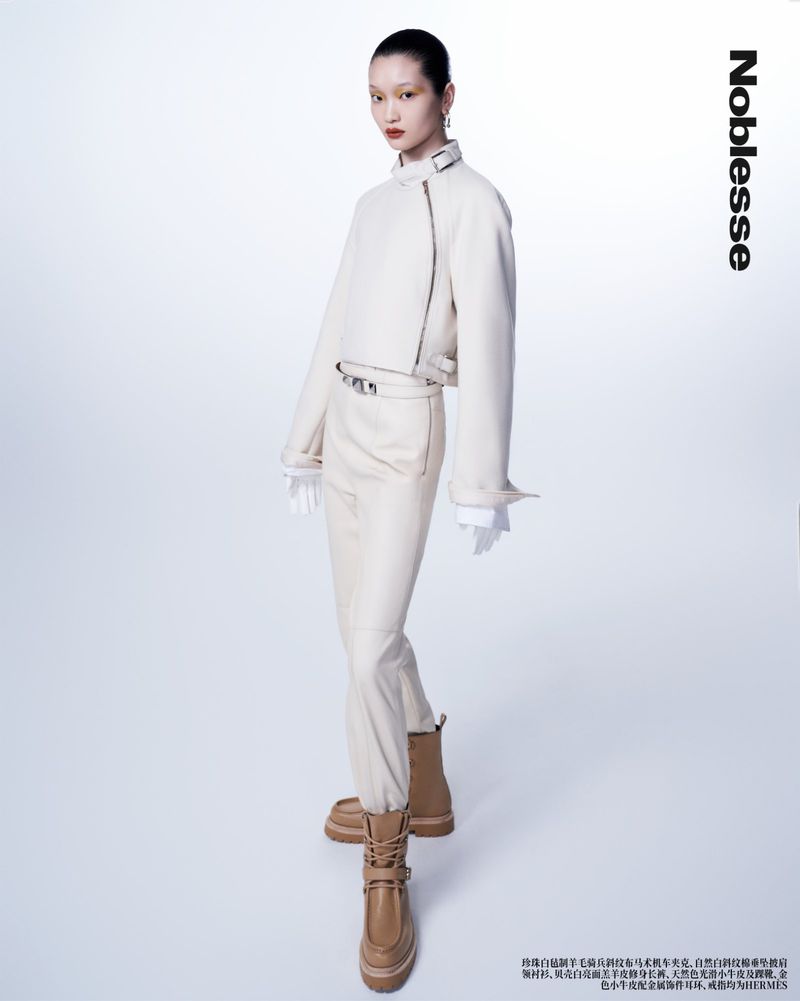 Editorial fashion shot of a woman posing in a cream-colored jacket and pants with tan boots against a white background in a studio.