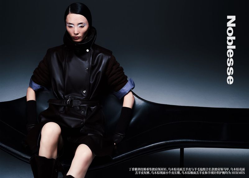 Editorial fashion shot of an adult female model sitting on a black leather lounge chair wearing a dark coat, black gloves, and a netted headpiece.