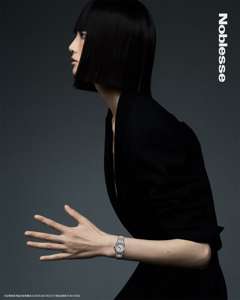 Woman in black jacket poses in a studio with silver watch. Black, blunt bob haircut, grey studio background. Fashion editorial shot.