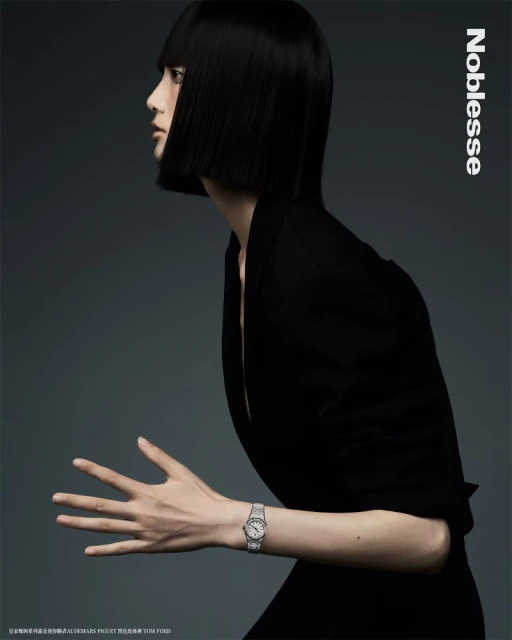 Woman in black jacket poses in a studio with silver watch. Black, blunt bob haircut, grey studio background. Fashion editorial shot.