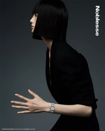 Woman in black jacket poses in a studio with silver watch. Black, blunt bob haircut, grey studio background. Fashion editorial shot.
