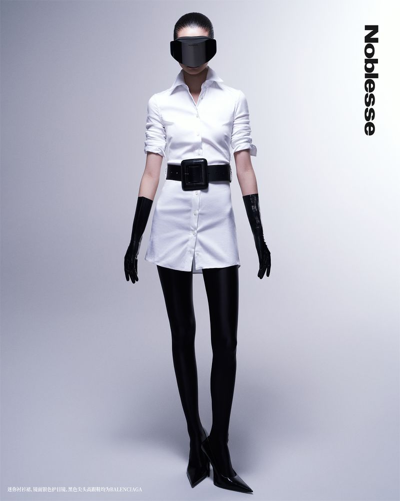 Full-length studio shot of a woman in a white shirtdress, wide black belt, black tights, gloves, and dark visor, against a light backdrop.
