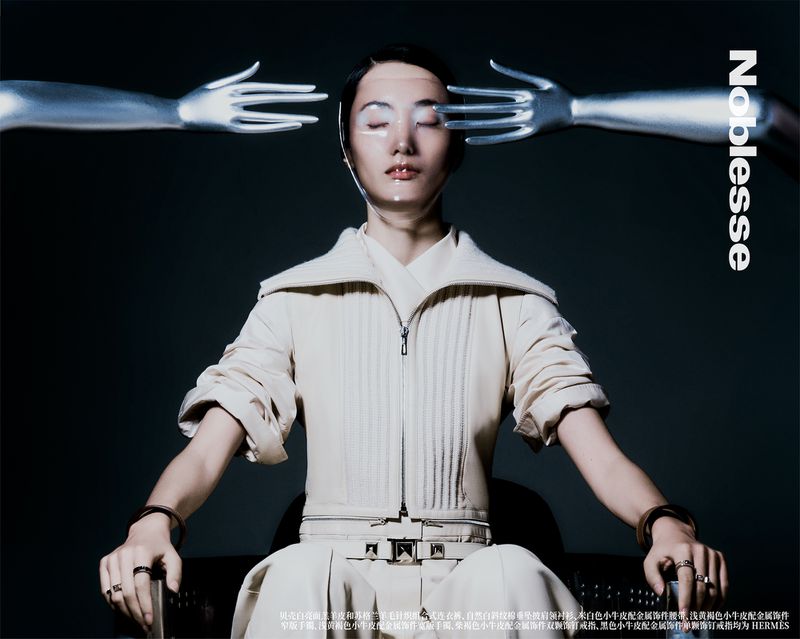 Asian female fashion model in avant-garde editorial-style pose wearing a beige suit, metallic makeup, and rings. Two silver hands frame face.